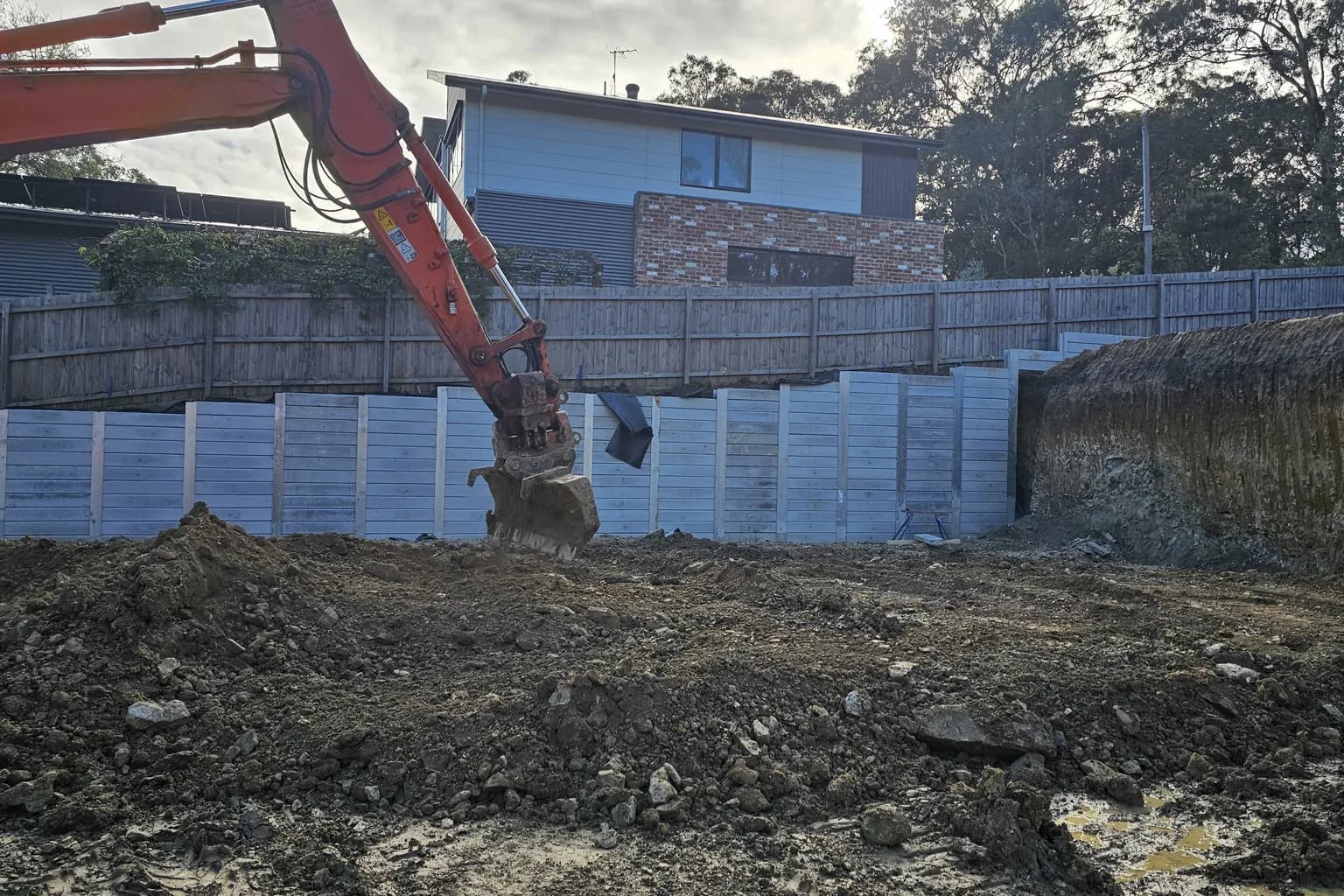 Excavator bucket in front of a stepped concrete sleeper retaining wall built along a boundary fence on a sloping block in Warrandyte